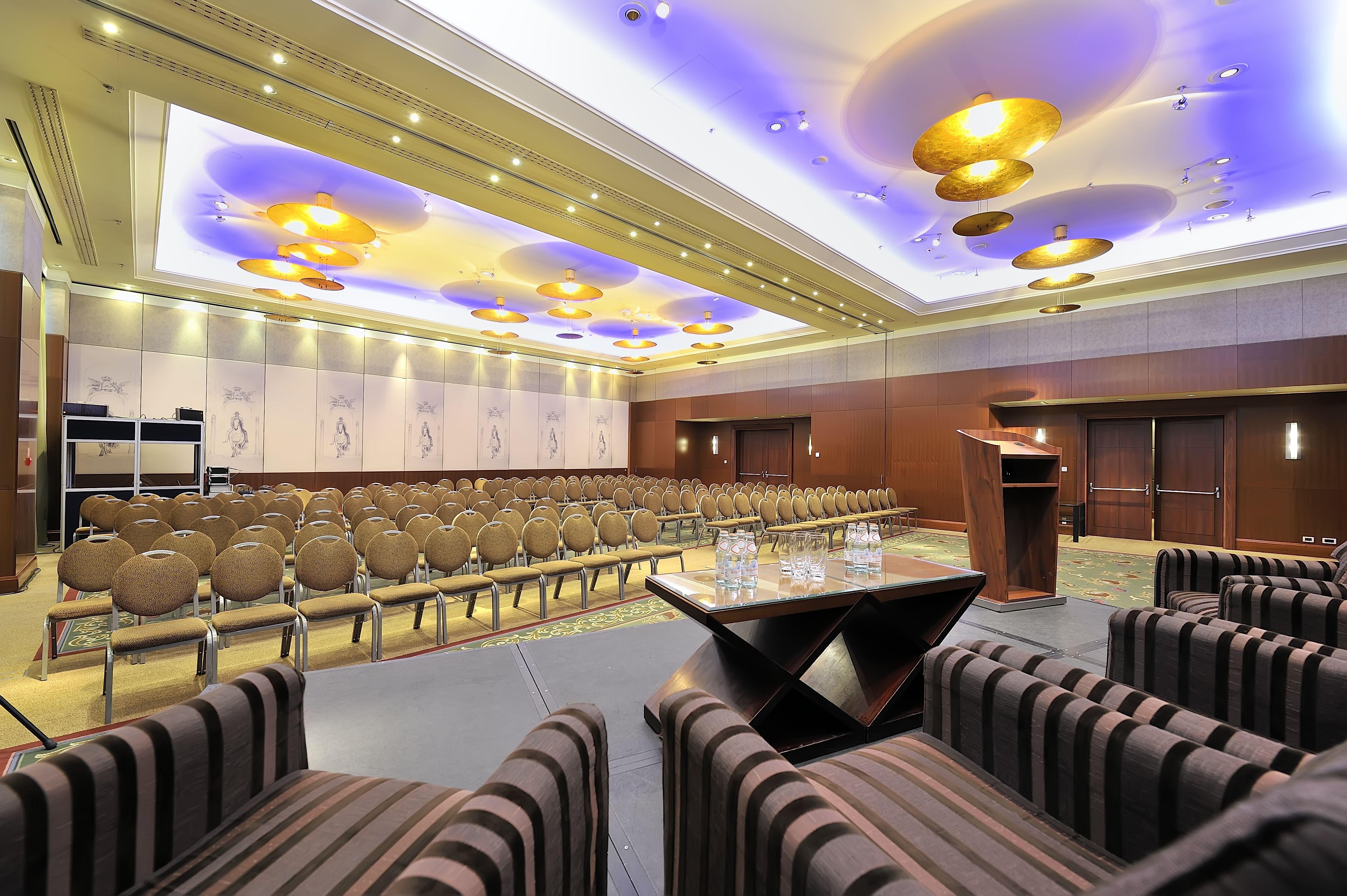 Regent Warsaw Hotel Exterior foto The photo shows a conference room or event space set up for a presentation or meeting. There are several rows of beige chairs arranged in front of a podium, which is positioned on a small stage. The room features elegant lighting, with decorative cei