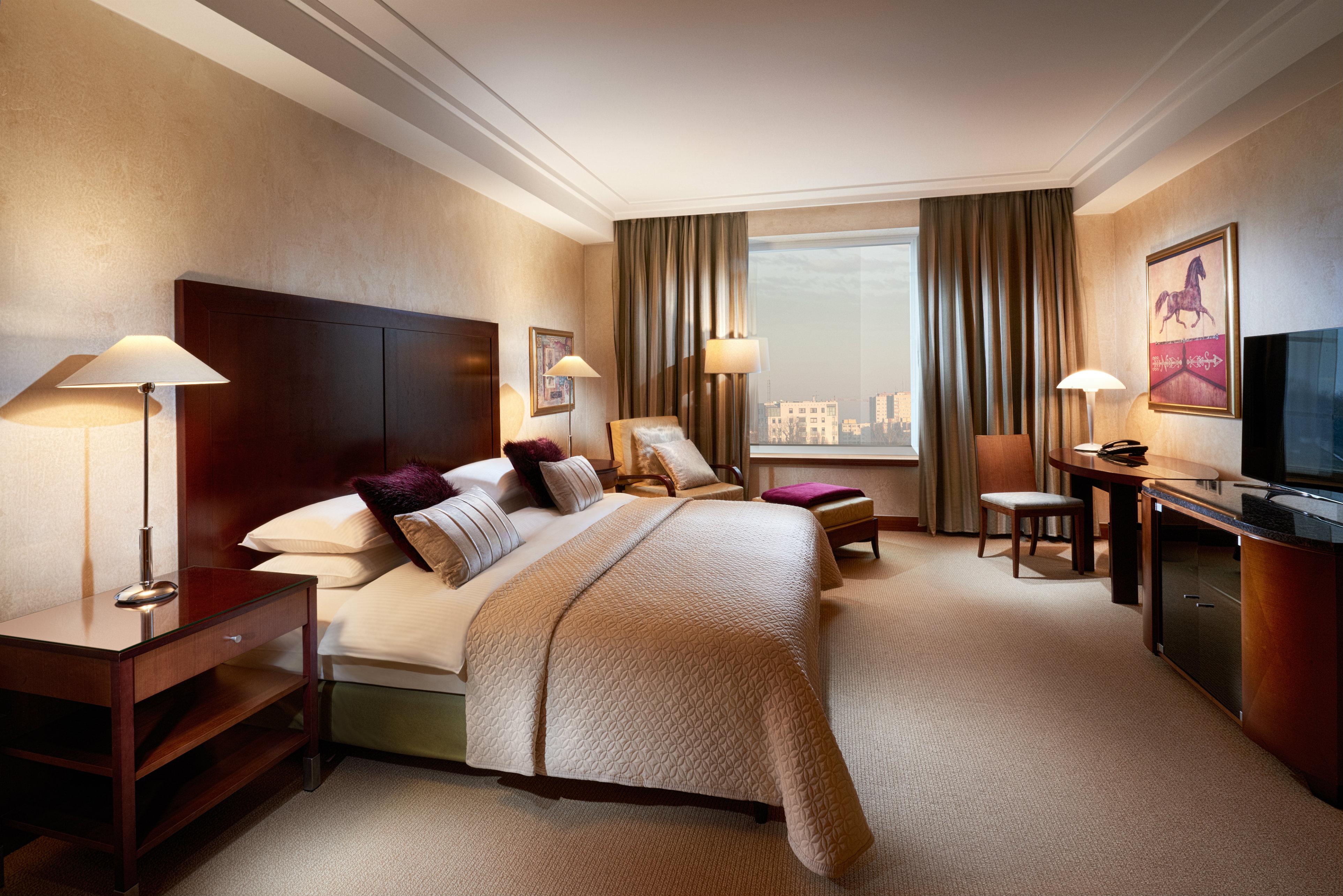 Regent Warsaw Hotel Exterior foto The photo shows a well-appointed hotel room featuring a large bed with white and cream bedding. There are bedside lamps on either side of the bed, and a comfortable armchair is positioned nearby. A window with long curtains allows natural light to en