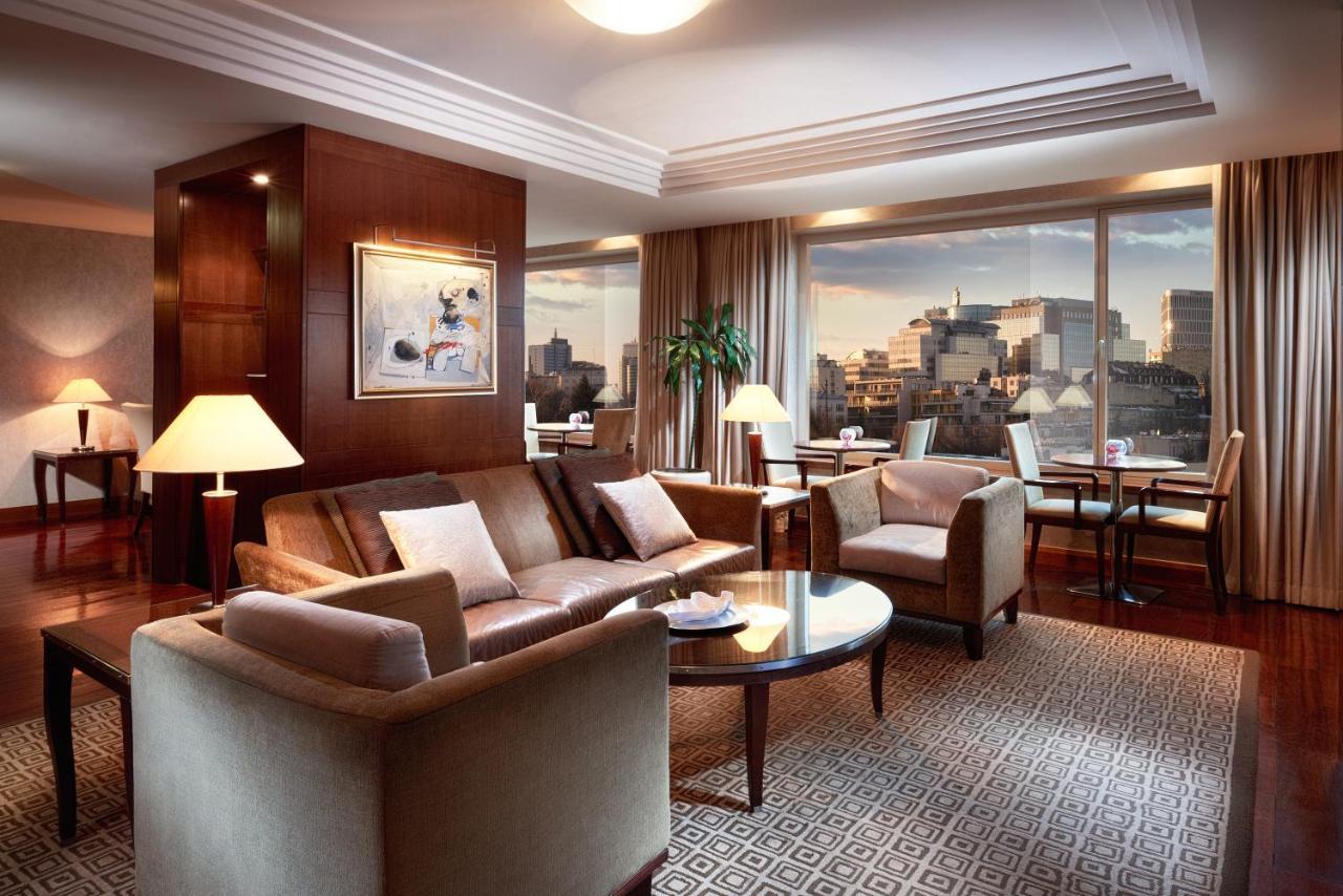 Regent Warsaw Hotel Exterior foto The image shows a stylish and modern living room with large windows offering a view of a city skyline. The room features a comfortable seating area with a sofa, armchairs, and a coffee table. There is a decorative piece of art on one wall, and soft l