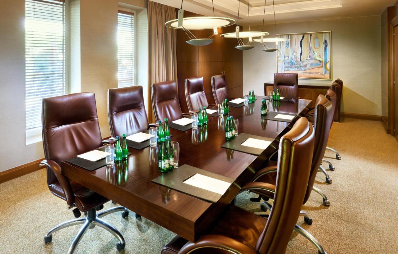Regent Warsaw Hotel Exterior foto The photo shows a modern conference room with a long wooden table surrounded by several brown leather executive chairs. On the table, there are glass bottles of water and paper notepads, suggesting preparation for a meeting. The room features large w